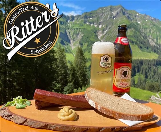 A plate with a sausage, fresh bread, mustard, and two beer bottles in front of an impressive mountain landscape. In the background, green hills and a clear blue sky can be seen.