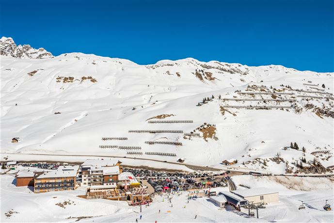 A snow-covered mountain landscape with a clear blue sky. In the foreground, there is a ski lift and a cozy restaurant with many visitors.