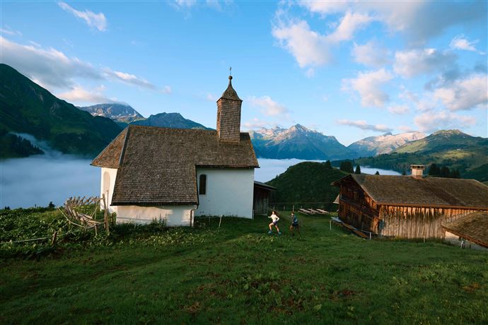 A picturesque church stands on a hill with mountains in the background. Gentle clouds surround the landscape, creating a peaceful atmosphere.