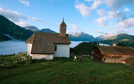 Eine malerische Kirche steht auf einem Hügel mit Bergen im Hintergrund. Sanfte Wolken umgeben die Landschaft und vermitteln eine friedliche Atmosphäre.