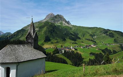Eine malerische Landschaft mit sanften Hügeln und einem Berg im Hintergrund. Im Vordergrund steht eine kleine Kirche, umgeben von grünen Wiesen.