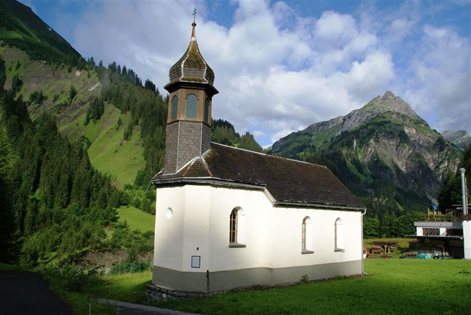 A small, charming church stands amidst green mountains. The sky is partly cloudy, and the landscape appears calm and idyllic.