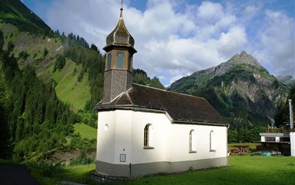 A small, charming church stands amidst green mountains. The sky is partly cloudy, and the landscape appears calm and idyllic.