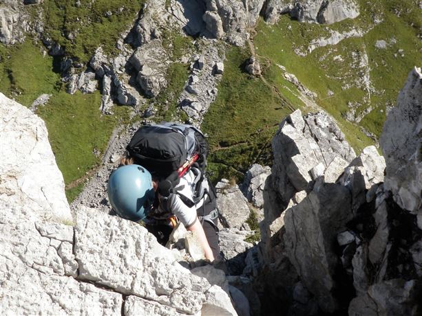 Een klimmer met een rugzak en een helm beklimt een steile rotswand. Op de achtergrond zijn groene heuvels en rotsachtige landschappen te zien.