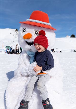A boy sits happily in the arms of a snowman in a snow-covered landscape. The sun is shining and the sky is blue.