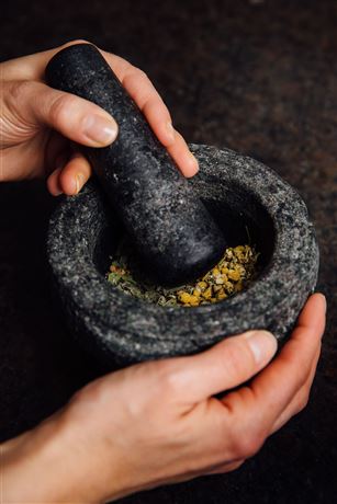 A person is using a mortar and pestle to grind herbs. The mortar is made of dark stone and contains colorful spices.
