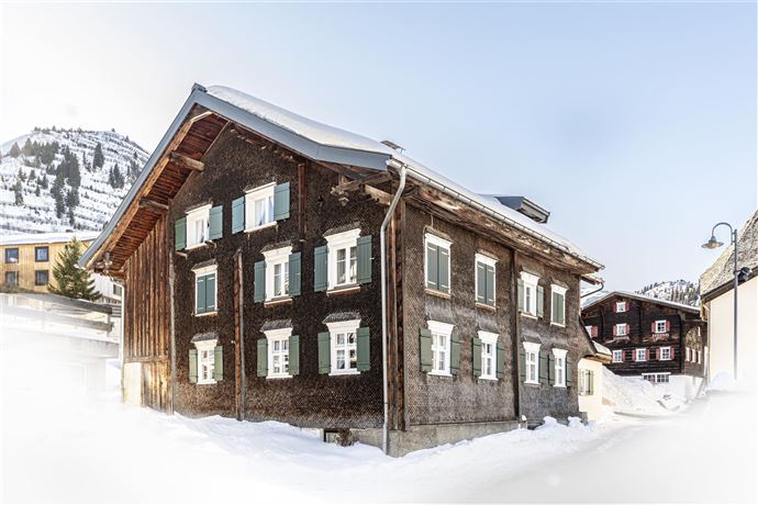 A traditional wooden house covered in snow in winter. In the background, snow-covered mountains and other buildings can be seen.