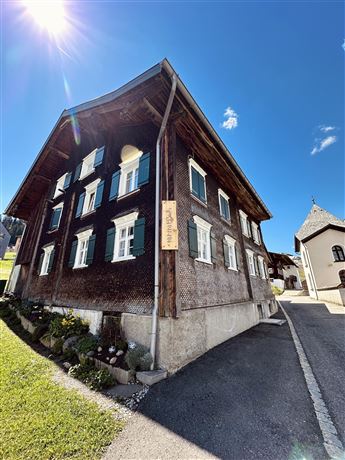 A traditional wooden house with green shutters and a clear blue sky. The surroundings are green and inviting.