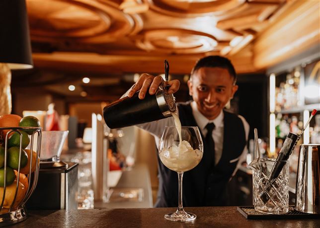 A bartender is pouring a cocktail into a glass. In the background, various drinks and a stylish bar are visible.