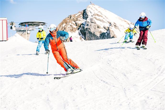Eine Gruppe von Skifahrern fährt auf einer verschneiten Piste. Im Hintergrund ist ein schneebedeckter Berg sichtbar.