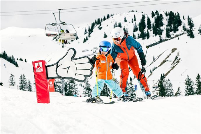 Ein Skilehrer hilft einem Kind beim Skifahren auf einer schneebedeckten Piste. Im Hintergrund sind Skilifte und verschneite Berge zu sehen.