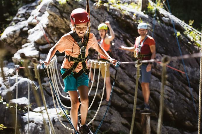 Een kind balanceert op een touw in de buitenlucht en draagt een helm. Op de achtergrond staan andere mensen die toekijken en applaudiseren.