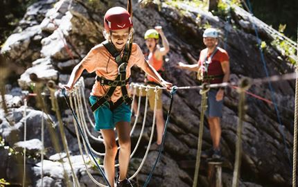 A child is balancing on a rope outdoors and wearing a helmet. In the background, there are other people watching and applauding.