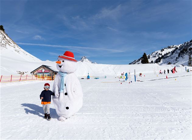 A snowman with a red hat stands in a snowy area holding the hand of a little boy. In the background, other children engaged in winter sports activities can be seen.