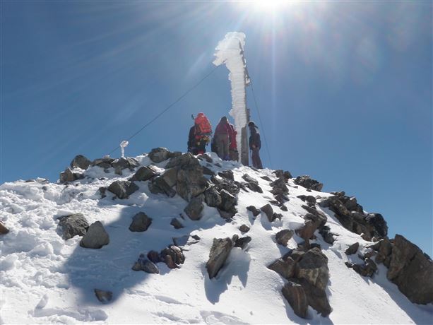 Een groep mensen staat op een met sneeuw bedekte top. Boven hen straalt de zon in een heldere blauwe lucht.