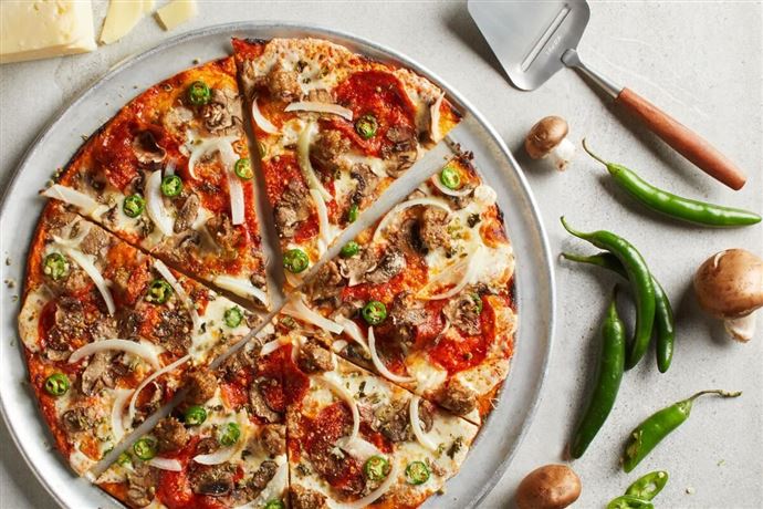 Eine frisch gebackene Pizza mit verschiedenen Belägen, darunter Salami, Zwiebeln und Jalapeños. Um die Pizza herum liegen frische Zutaten wie grüne Chilischoten und Champignons.