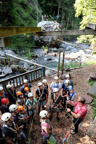 Een groep personen in helmen en klimmateriaal luistert naar een instructeur in een avonturenpark. Op de achtergrond zijn bomen en een stromende rivier zichtbaar.