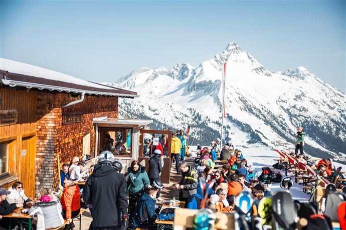 A bustling ski lodge area with many people and ski equipment. In the background, impressive snow-covered mountains and a clear blue sky can be seen.