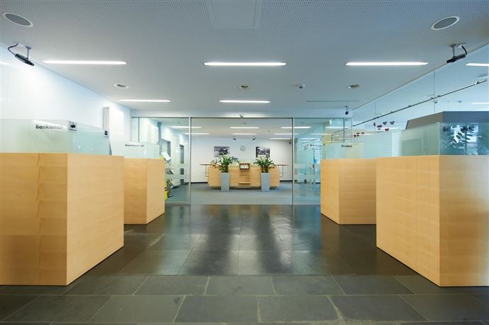 A modern office with glass partition walls and wooden-colored reception desks. The room is bright and well-lit, with plants for decoration.