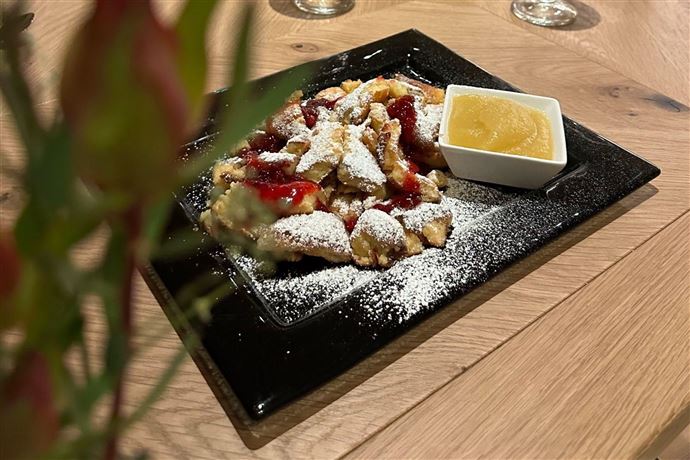 An appetizing dessert on a black plate, consisting of pieces of Kaiserschmarrn with powdered sugar and a portion of apple sauce. Next to it, a stylish decoration made from a plant is visible.