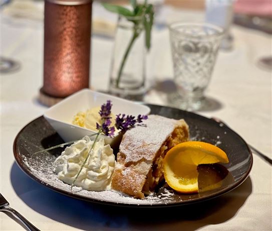 A served apple strudel on a black plate, garnished with cream and fresh oranges. In the background stands a decorative candle and a glass of water.