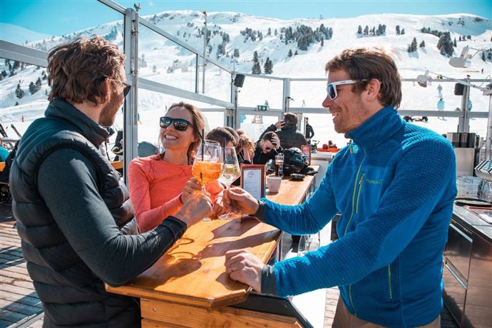 Three people are enjoying drinks on a sunny terrace in the snow. In the background, skiers and snowy mountains can be seen.
