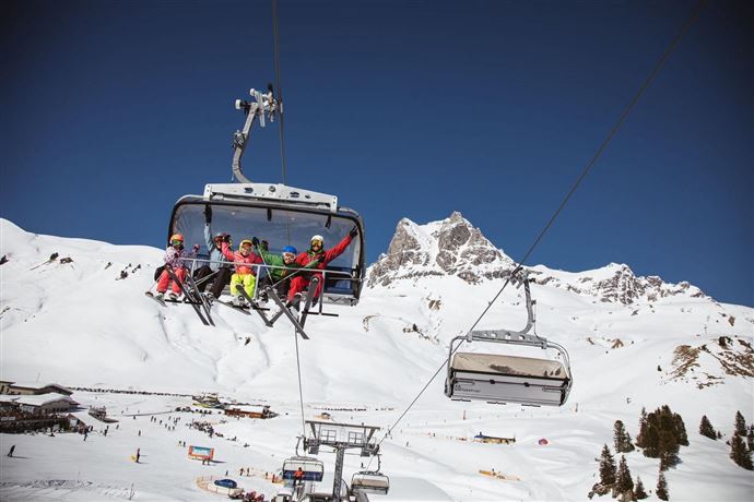 Eine Skiliftfahrt mit mehreren Personen in bunten Skianzügen. Im Hintergrund sind schneebedeckte Berge und ein klarer blauer Himmel zu sehen.