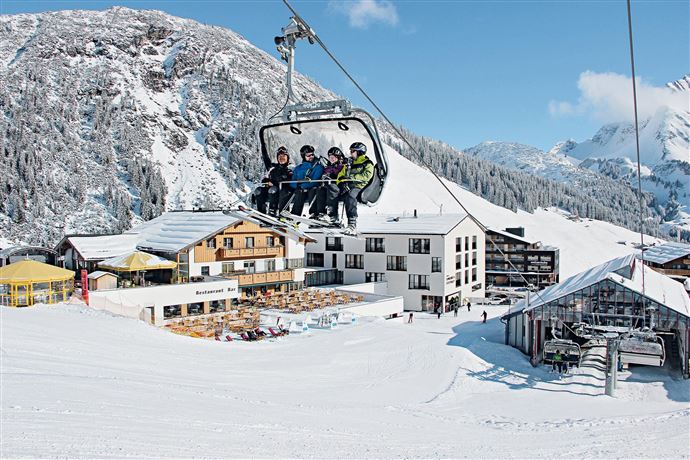 A ski lift ride with several people in the midst of a snowy winter landscape. In the background are mountains and a mountain hut.