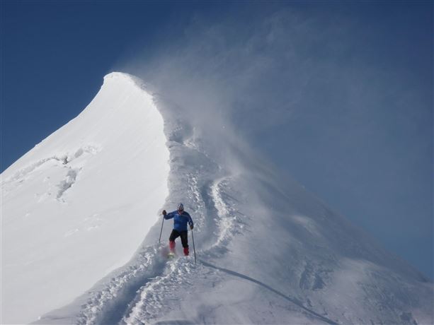 Een skiër beklimt een met sneeuw bedekte top. De lucht is helder en er waait een lichte wind.