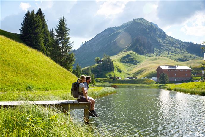 A hiker sits by the shore of a calm lake, surrounded by green hills and mountains. The landscape is sunny and idyllic, perfect for outdoor activities.