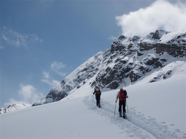 Two hikers are walking through snowy landscapes in the mountains. The sky is clear with some clouds, and the mountain peaks are impressive.