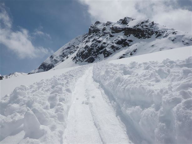 A snow-covered mountain with a clear sky. A trail in the snow leads to the summit region.