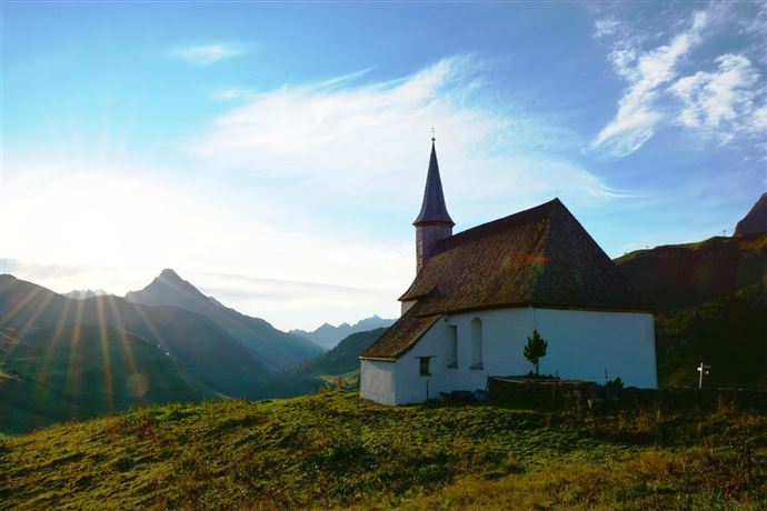 Eine malerische Kirche auf einem Hügel, umgeben von Bergen. Im Hintergrund strahlt die Sonne und sorgt für eine wunderschöne Atmosphäre.