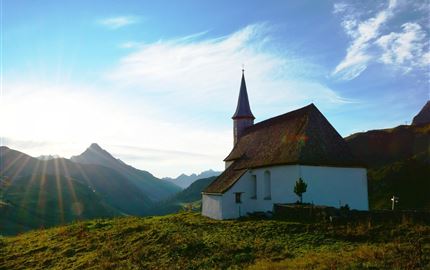 Eine malerische Kirche auf einem Hügel, umgeben von Bergen. Im Hintergrund strahlt die Sonne und sorgt für eine wunderschöne Atmosphäre.