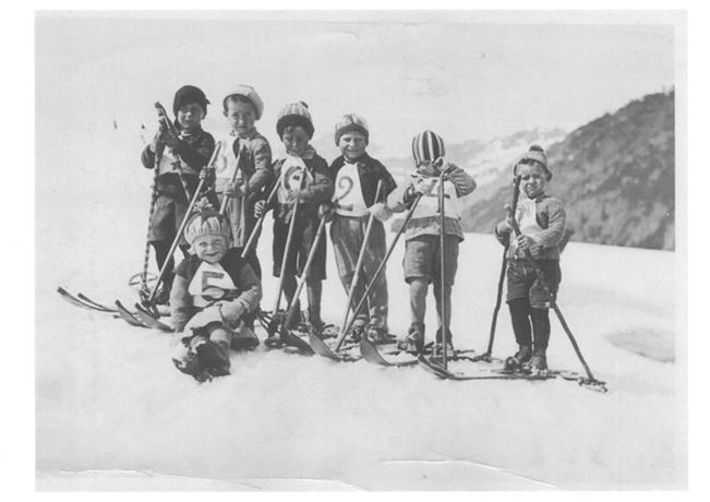 A group of eight children stands on skis in the snow. They are smiling and wearing winter sports clothing in a mountainous environment.