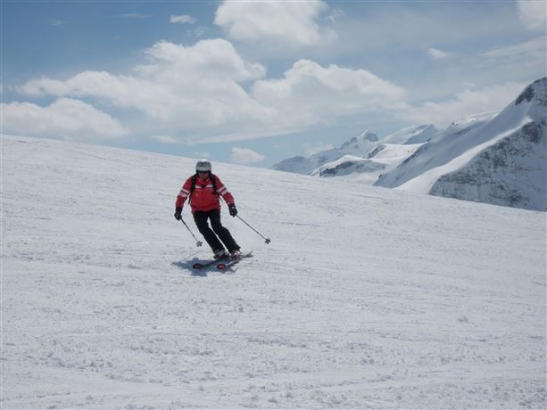 A skier is skiing down a snow-covered slope. In the background, there are tall mountains and a blue sky visible.