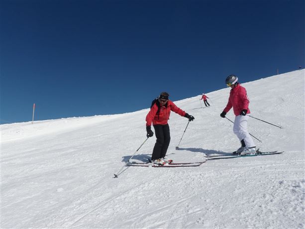 Two skiers on a snowy slope under a clear blue sky. In the background, another skier can be seen.