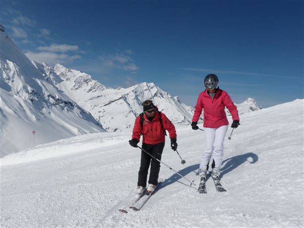 Two skiers on a snowy slope in the mountains. In the background, there are high, snow-covered peaks and a blue sky.