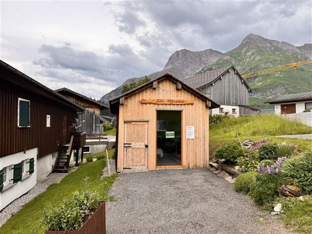 A wooden building with an inviting front door stands amidst green meadows and mountains. The clouds in the sky give the scene a tranquil atmosphere.