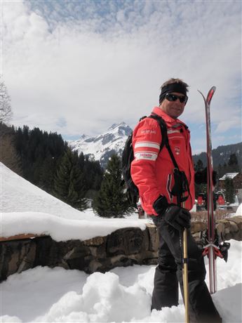 A skier stands in the snow with mountains in the background. The sky is clear and the landscape is wintry.