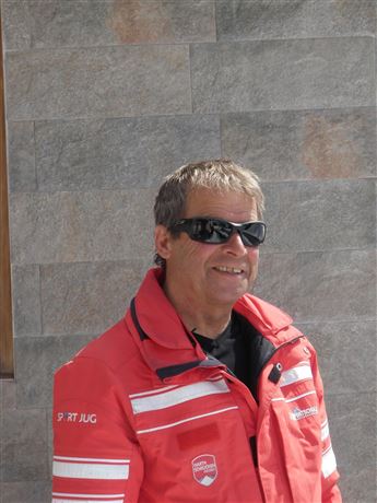 A smiling man in a red jacket stands in front of a gray wall. He is wearing sunglasses and looks friendly.