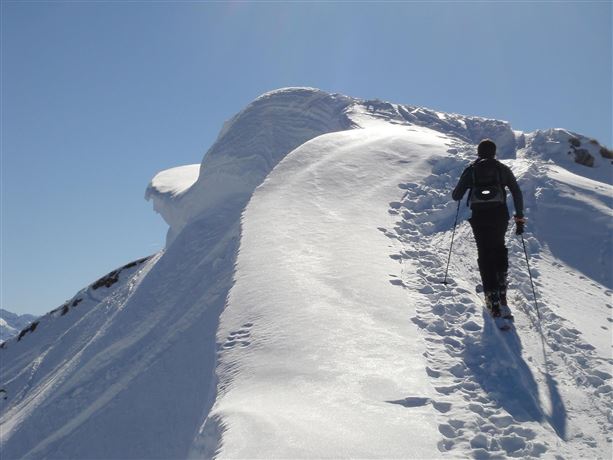 A hiker climbs a snow-covered peak under a clear sky. The edge of the mountain offers an impressive view.