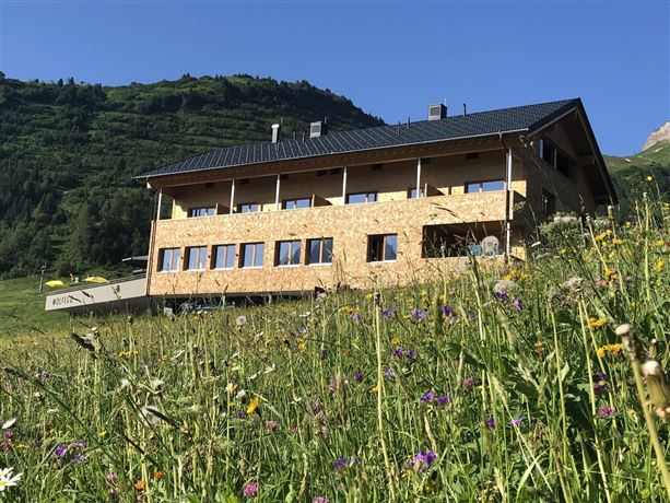 A modern wooden house stands in a meadow with colorful flowers. In the background, there are green hills and a clear blue sky.