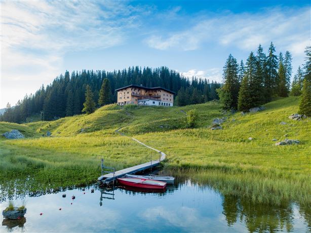 A picturesque lake surrounded by lush greenery and tall trees. In the background, there is a charming building on a gentle hill.