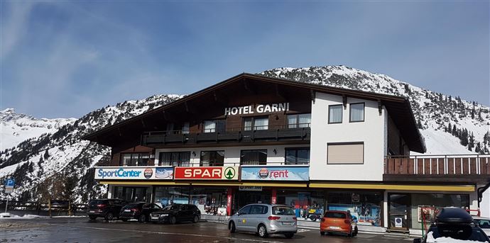 A hotel named "Hotel Garni" in the mountains, surrounded by snow-covered peaks.  
On the ground floor, there are shops such as a sports center and a supermarket.