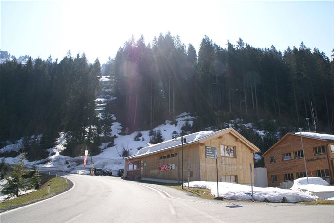 A winding road leads through a wintry landscape with snow-covered areas. In the foreground, wooden buildings stand surrounded by dense coniferous trees.