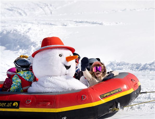 A cheerful snowman with a red hat is riding on a rubber boat in the snow. The group of children is having fun and enjoying the winter scenery.
