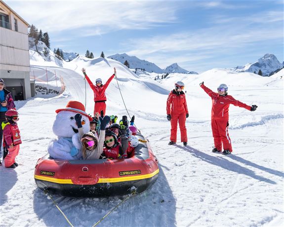 A group of children sits in a red inflatable boat, surrounded by snow. In the background, ski instructors in red suits and a snowman with a hat stand.
