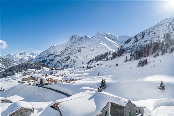 A winter landscape with snow-covered mountains and a small village below. The sky is clear and blue, highlighting the winter beauty.