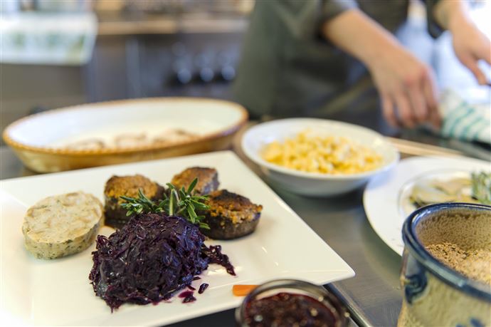 A plate with delicious food, consisting of meat, red cabbage, and side dishes. In the background, a person can be seen preparing more dishes.